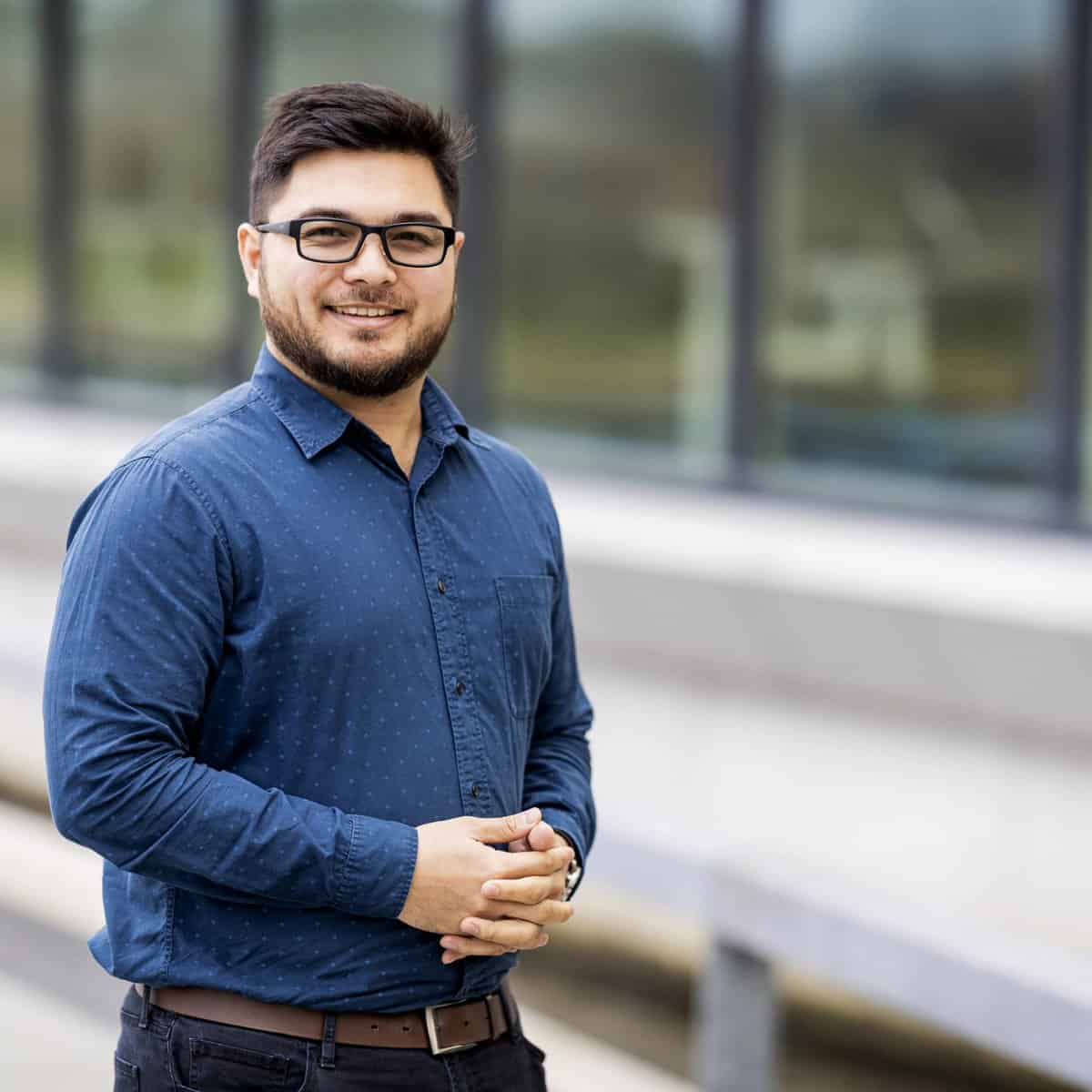 Pinoy scientist Jowi Miranda has dark hair and beard, wearing glasses and a blue button-up shirt, stands outdoors in front of a building with large windows. He is smiling and clasping his hands together.