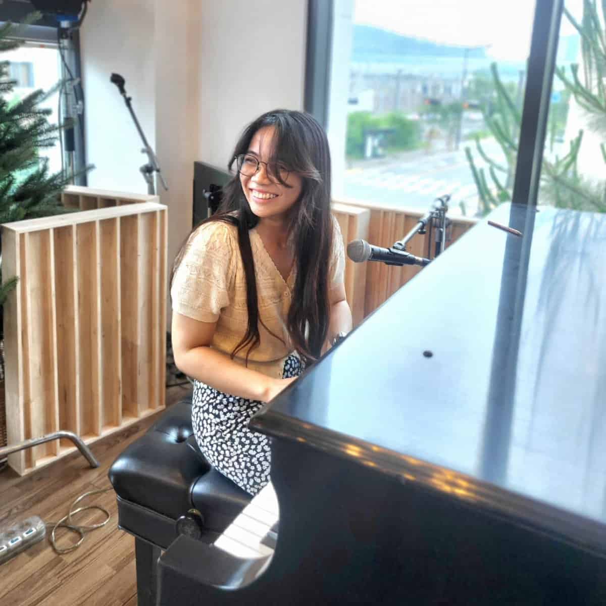 May Angelu Madarang is playing a piano with a mic in front of her. She is looking at the audience with a big smile.