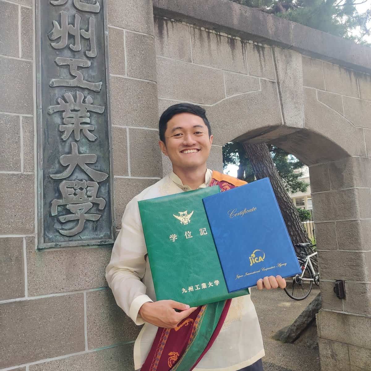 John Paul Almonte is dressed in barong and sablay as he stands while holding his certificate from JICA.
