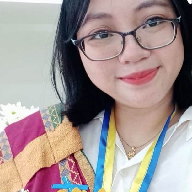 Ma. Criselda Baldago has glasses and black hair. She is smiling, wearing a white shirt and sablay, and a blue and yellow ribbon around her neck.
