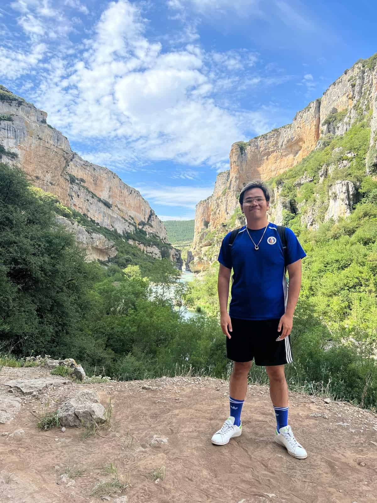 Cesar Miguel Fabro is standing on a dirt path with a backpack. Behind him is a scenic view of a lush green valley and towering cliffs under a blue sky with scattered clouds.