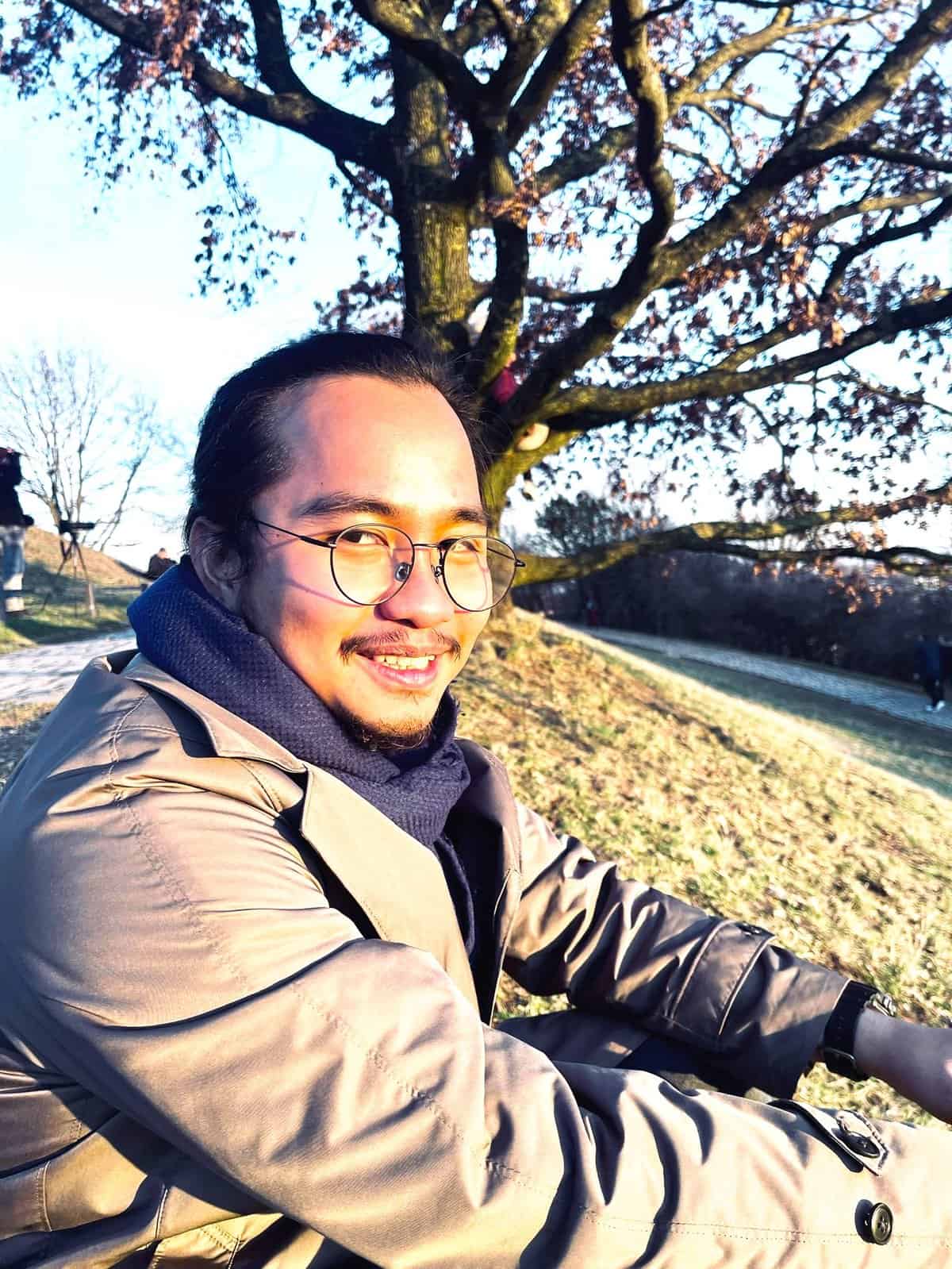 Jason Sebastian Simon is wearing glasses and a smile. He is sitting outdoors on a grassy hill. He is wearing a brown coat and a dark scarf.