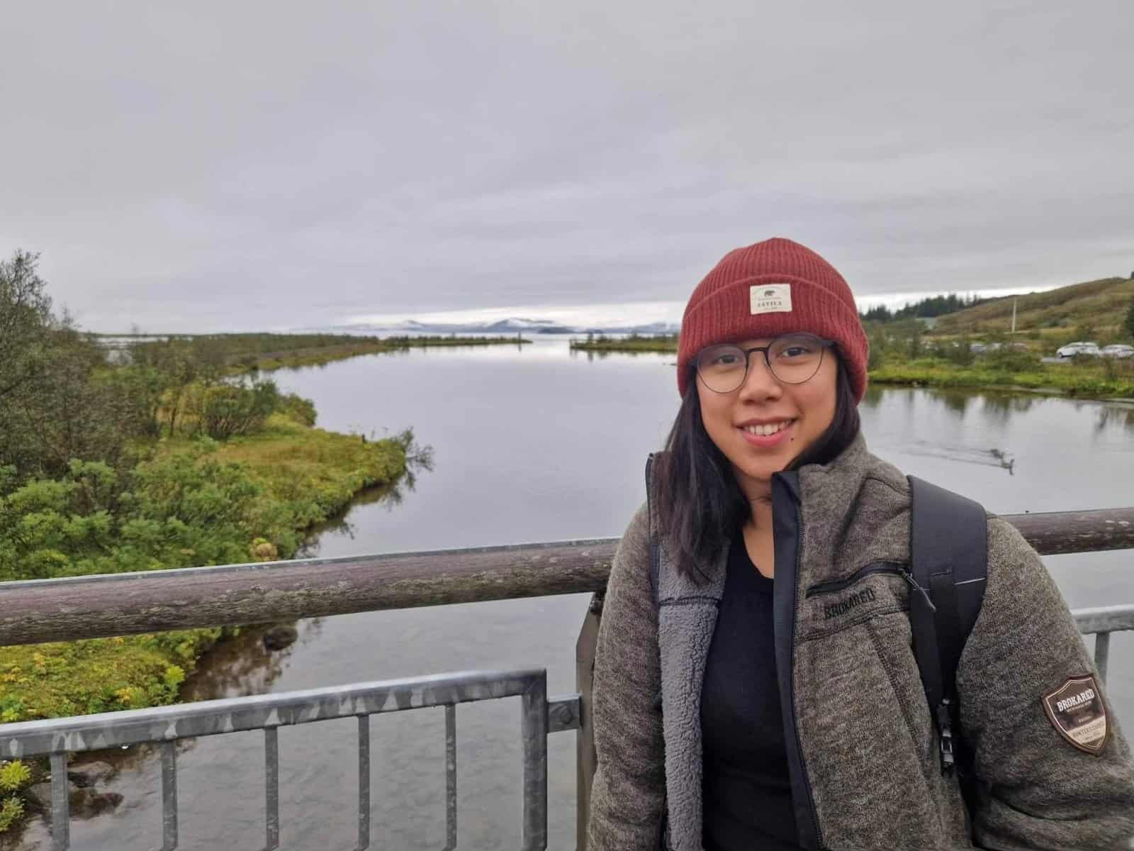 Kimberly Lagrana is wearing a red beanie, glasses, and a green jacket with a backpack. She stands on a bridge over a serene river with surrounding greenery