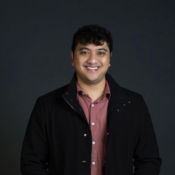 Jason Paolo Telles has short curly hair. He is smiling at the camera and wearing a black jacket over a maroon shirt.
