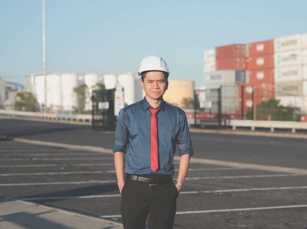 Jacob is wearing a white hard hat, blue shirt, and red tie. He is standing in an industrial area with shipping containers and metal tanks in the background.