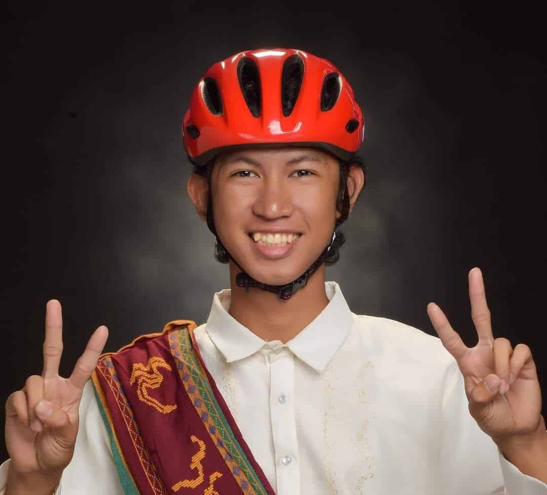 Pinoy scientist Ramir Angeles wearing a red cycling helmet with his barong and sablay, poses with both hands making peace signs.