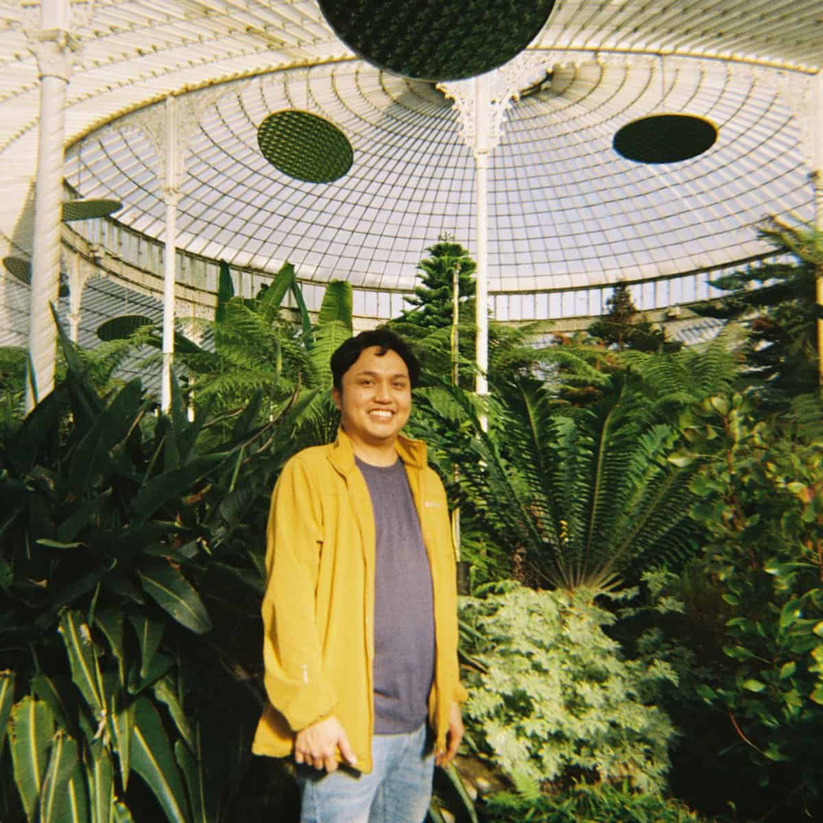 Roberth Rondilla is in a yellow jacket, standing smiling in a lush greenhouse filled with ferns and plants.