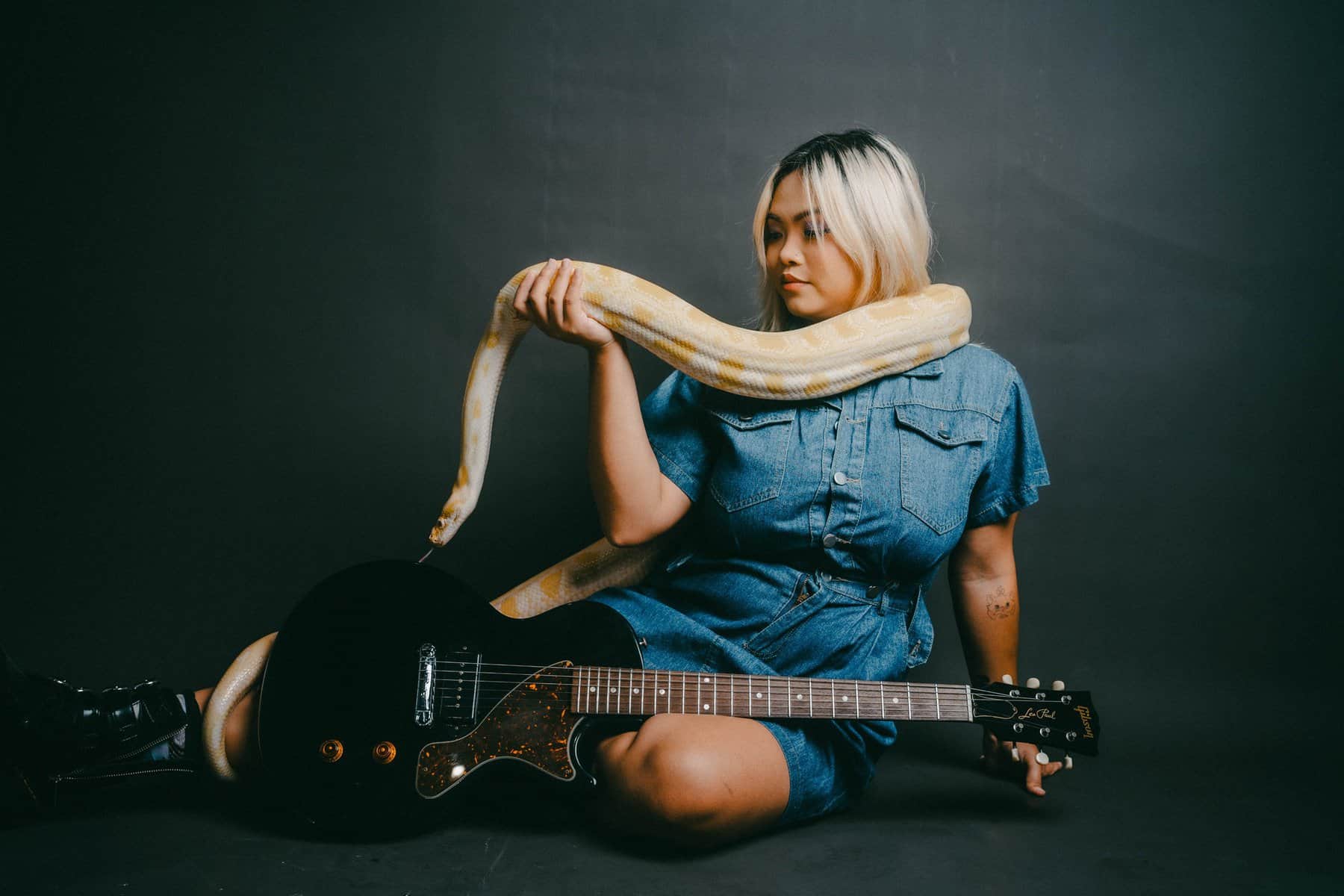 Jazz Ong sits on the floor in a dark room, holding a yellow snake draped over their shoulders. She is wearing a denim dress and black boots, with a black electric guitar resting in front.