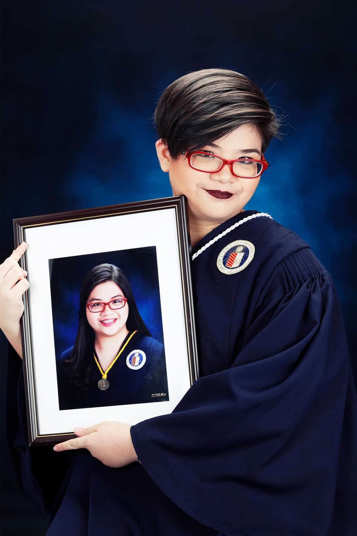 Becca Yu is wearing a navy graduation gown and red glasses. She holds a framed photo of themselves in a similar outfit.