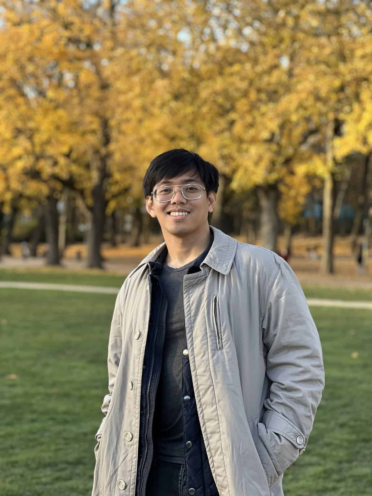 Rhowell Tiozon is wearing glasses, standing on a grass field before trees with autumn foliage. He is wearing a light-colored coat over dark clothing.