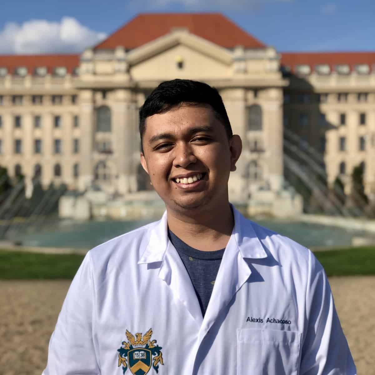 Pinoy Scientist Alexis Achacoso is in a white lab coat, stands smiling in front of a large, ornate building with a red roof and fountains.