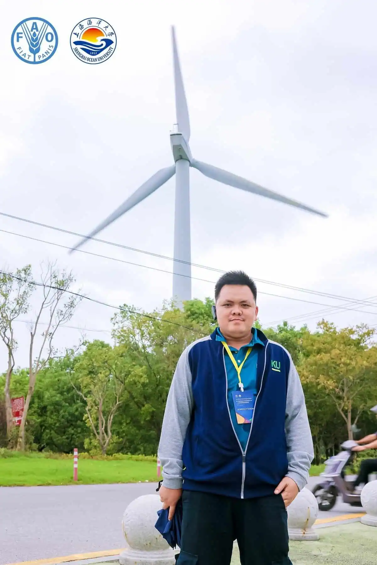 Pinoy scientist Jasper Kit Tangal stands outdoors in front of a large wind turbine, wearing a navy jacket with a lanyard badge.
