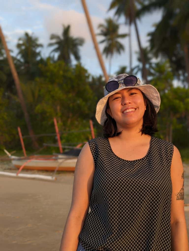 Pinoy scientist Gabrielle Leung wearing a sun hat, sunglasses, and a sleeveless top, smiles on a tropical beach with palm trees and a boat in the background, bathed in warm sunlight.