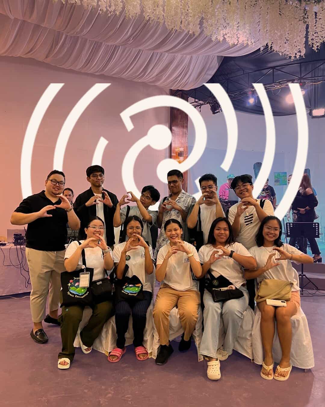 Members of the D&C Group pose together indoors, some sitting and some standing, making heart shapes with their hands.