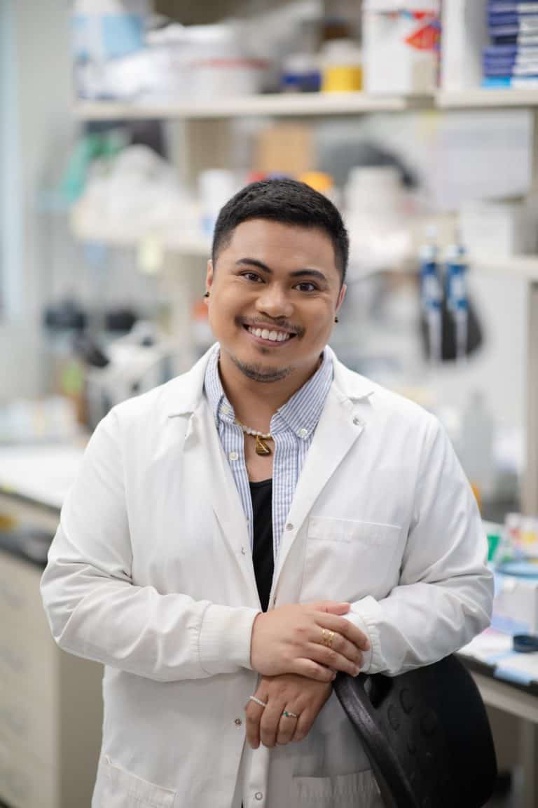 Pinoy Scientist Angelico “Anjo” Obille in a white lab coat standing in a laboratory, with shelves of lab equipment and supplies in the background.