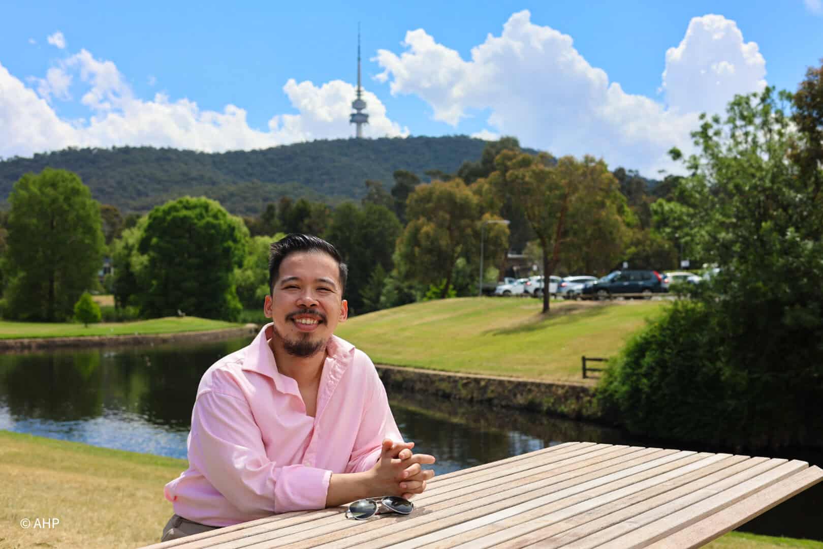 Pinoy scientist Aron Harold Pamoso is in a pink shirt, sits at a wooden picnic table by a pond, smiling at the camera.
