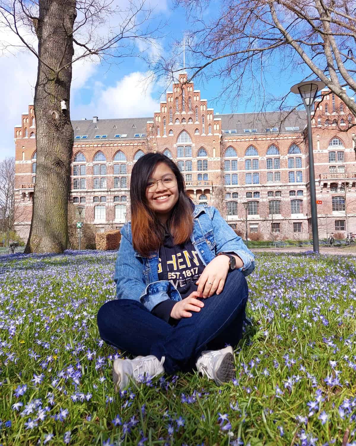 Pinoy scientist Gizelle Danine Jamero has long hair, glasses, and a denim jacket sitting smiling on grass covered with small purple flowers, with a large brick building and leafless trees in the background on a sunny day.