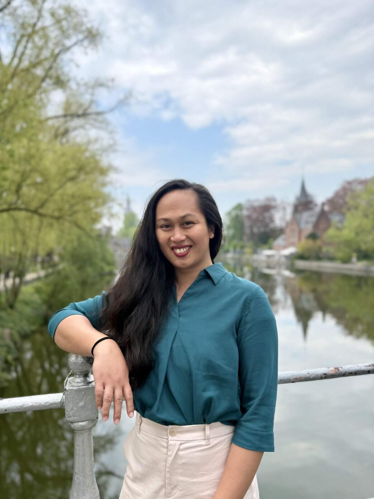 Pinoy scientist Joey Cabasan with long dark hair, wearing a teal blouse and beige pants, smiles while standing on a bridge with greenery, water, and historic buildings in the background under a cloudy sky.