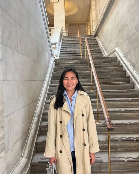 Pinoy scientist Nicole Ortiz has long dark hair, wearing a beige trench coat and blue shirt, smiles while standing in front of a grand marble staircase with ornate walls and a handrail.