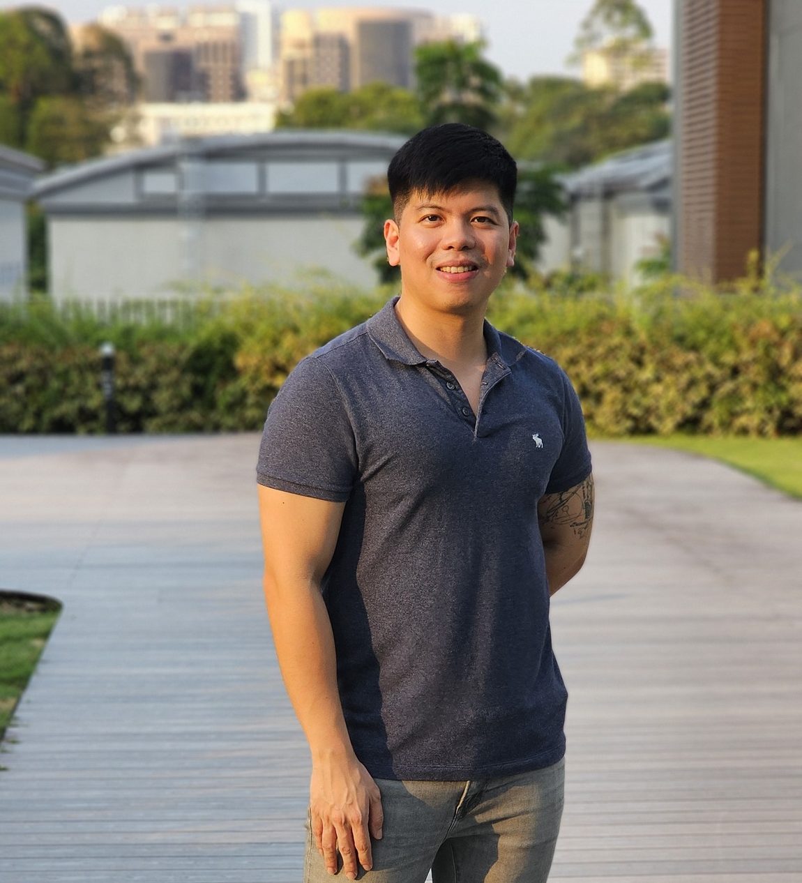 Pinoy scientist Ryan De Sotto wears a navy blue polo shirt, gray pants, and white sneakers, stands outdoors on a wooden walkway, smiling at the camera.