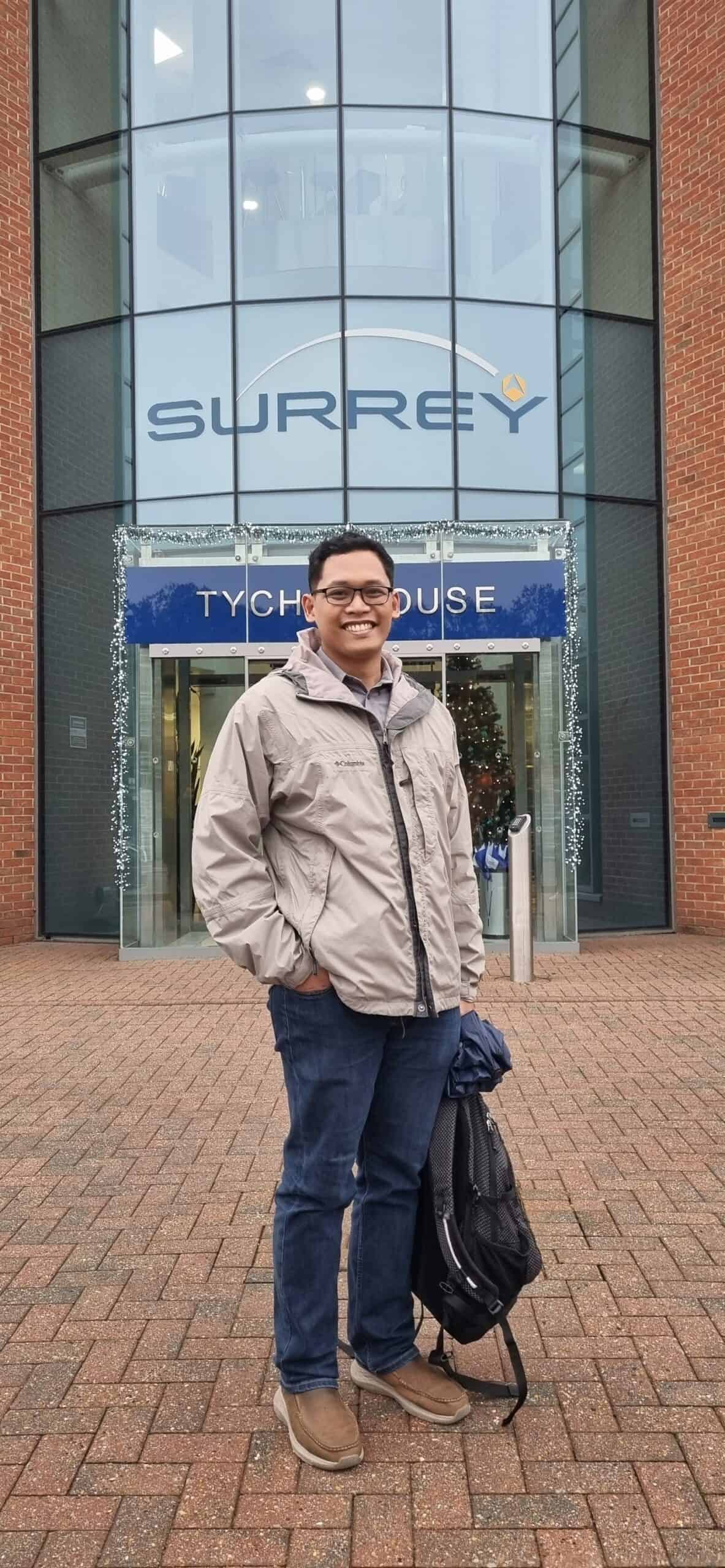Pinoy scientist Izrael Zenar Bautista is wearing glasses, a light gray jacket, and blue jeans stands smiling in front of a building entrance