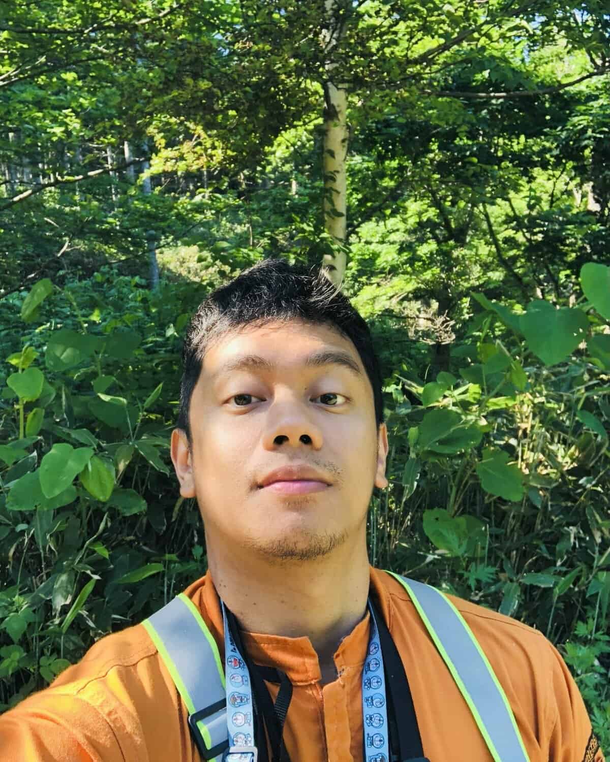 Pinoy Scientist James Cesar Refran is wearing an orange shirt and a reflective vest in a selfie outdoors, standing in front of lush green foliage and trees on a sunny day.
