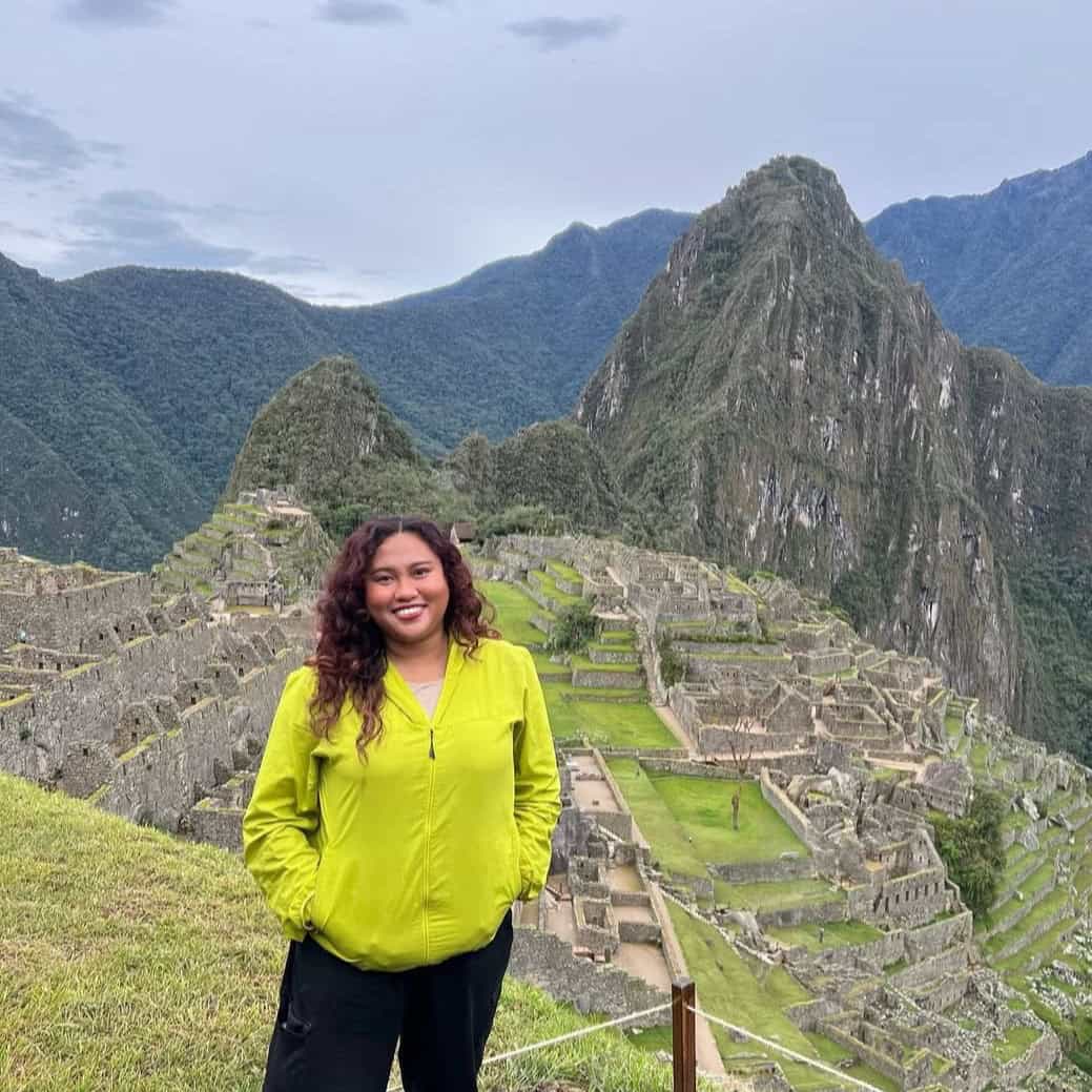 Pinoy scientist Cristal Balaga is wearing a bright yellow jacket, smiling in front of the ancient ruins of Machu Picchu.