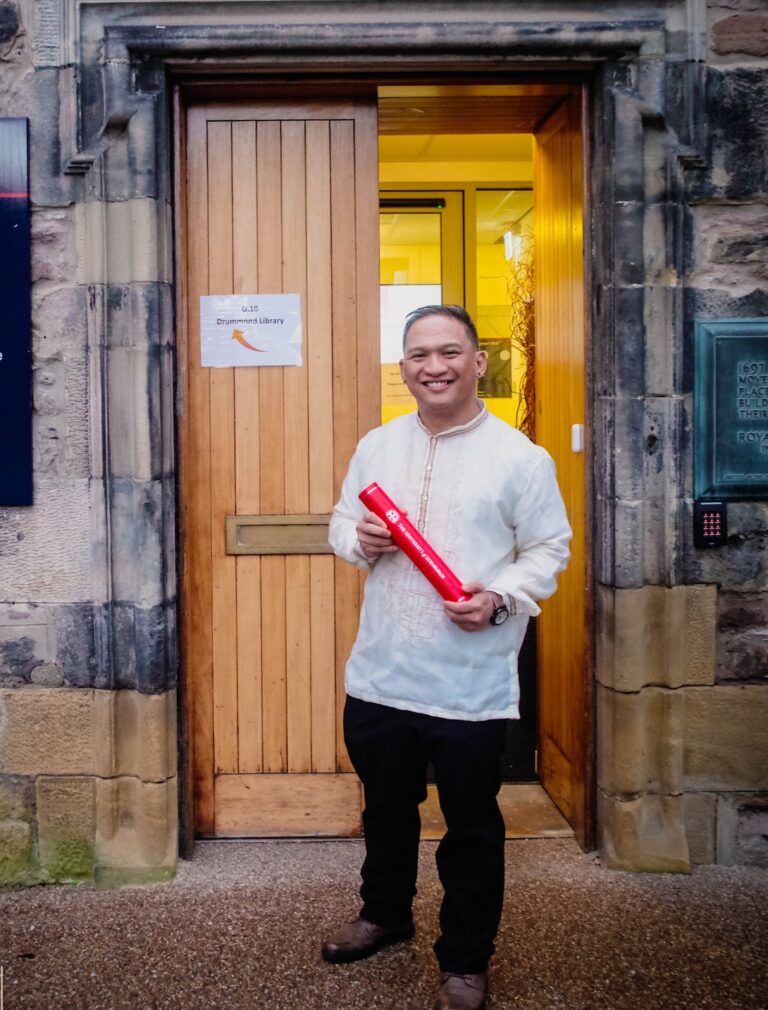 Pinoy scientist JJ Gepanaga in a barong holds a red diploma tube while standing in front of an open doorway at Old Surgeons’ Hall, University of Edinburgh