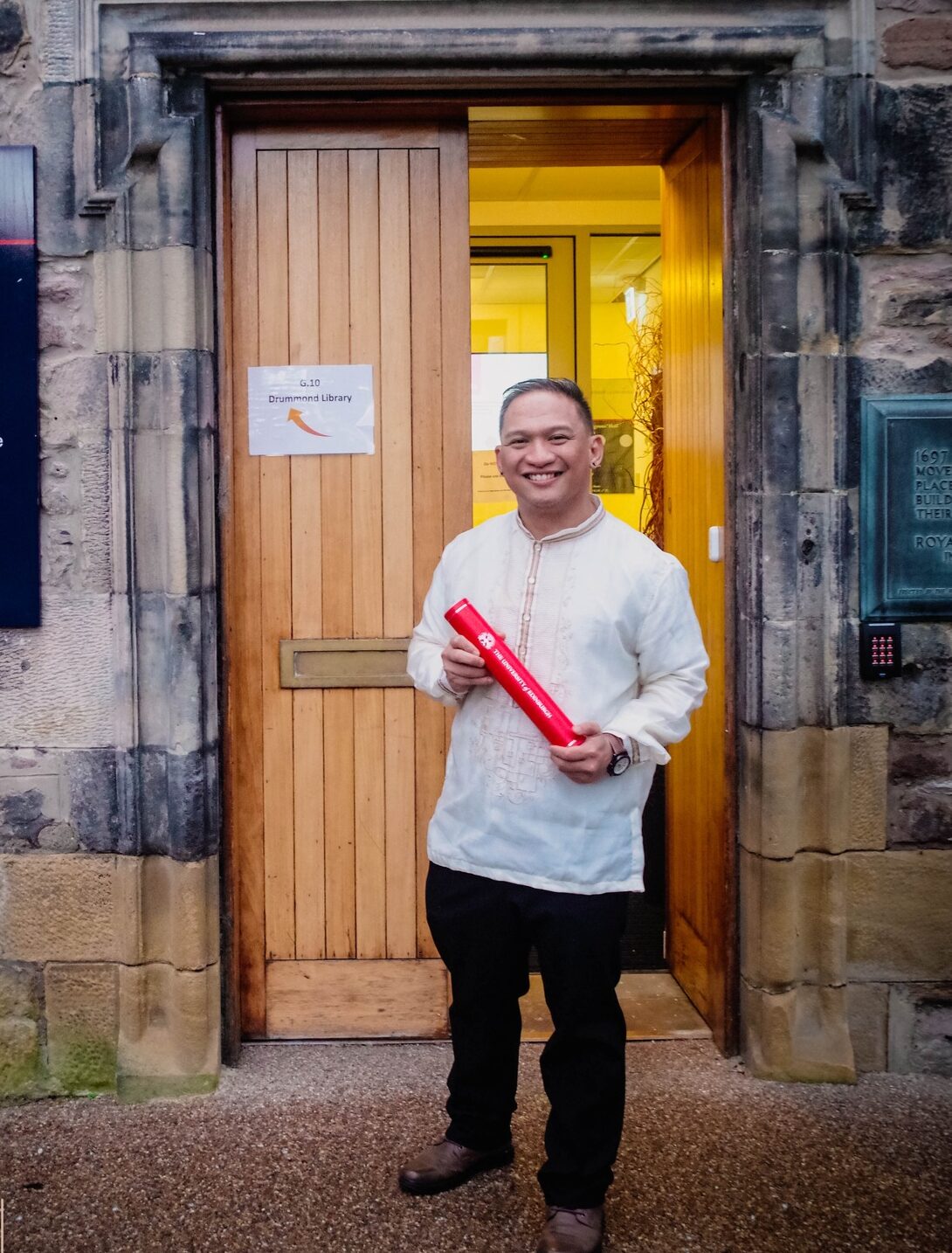Pinoy scientist JJ Gepanaga in a barong holds a red diploma tube while standing in front of an open doorway at Old Surgeons’ Hall, University of Edinburgh