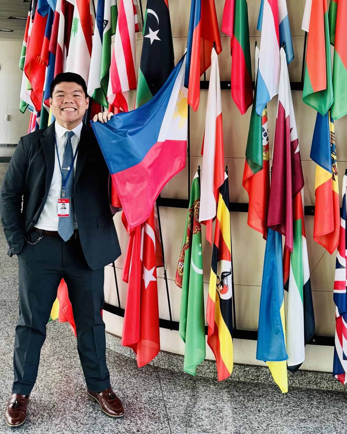 Pinoy scientist Dr. Angel Bautista VII in a suit and tie smiles while holding the Philippine flag among a display of various international flags indoors.