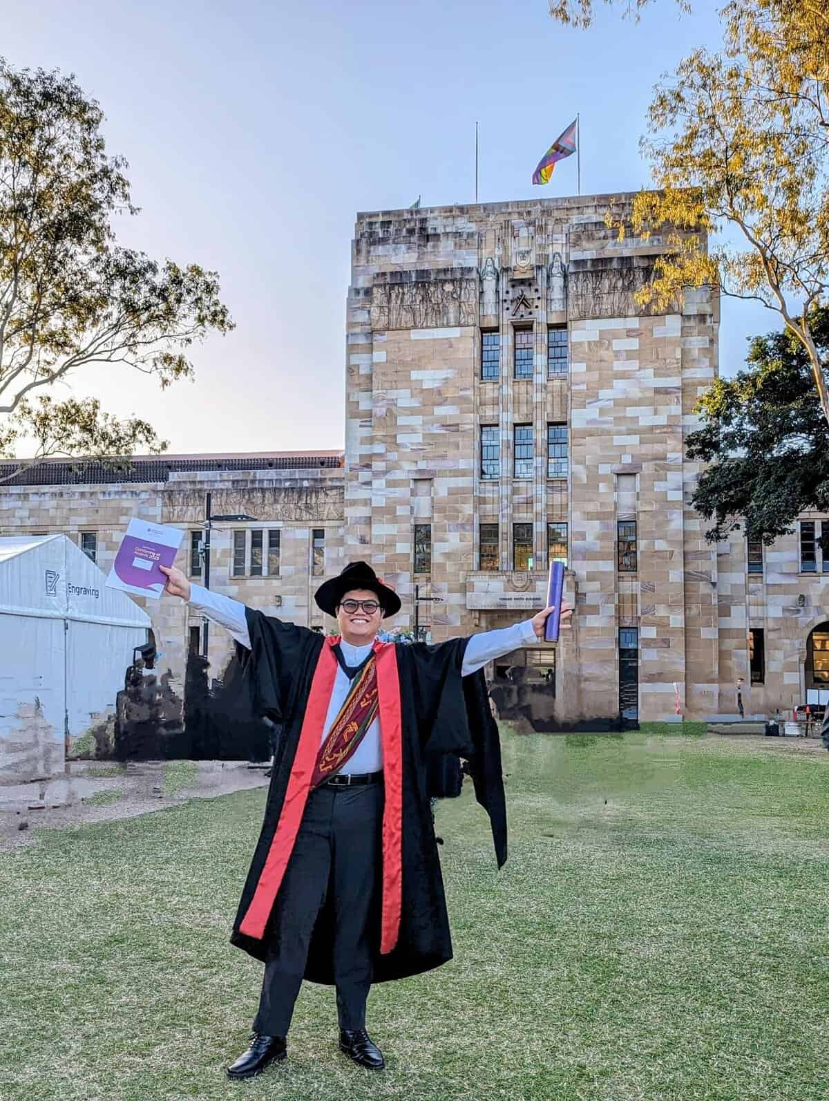 Pinoy scientist Mario Jr Rebosura in a graduation cap and gown standing on grass with arms raised, holding certificates, in front of a stone university building.