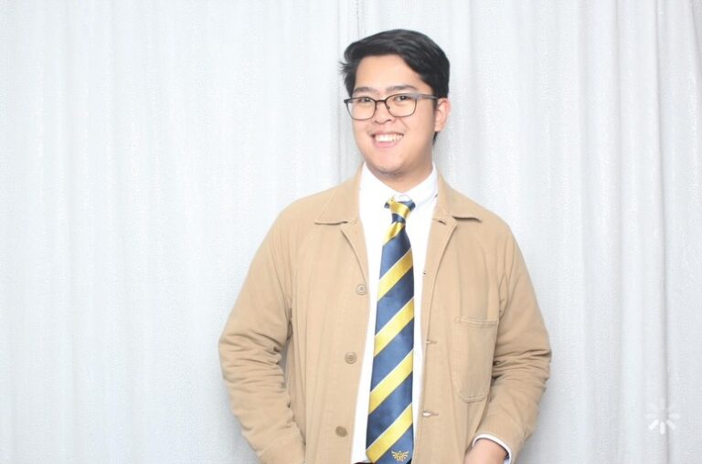 Pinoy scientist Aaron Gabriel Espinosa wearing glasses, a tan jacket, white shirt, and a blue and yellow striped tie stands smiling in front of a white curtain background with one hand in his pocket.