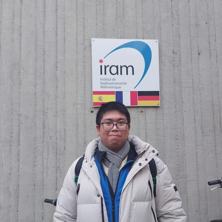 Pinoy scientist Daryl Joe Santos is wearing glasses, a white winter jacket, and a scarf. He stands in front of a wall with a sign for IRAM (Institut de Radioastronomie Millim&eacute;trique).