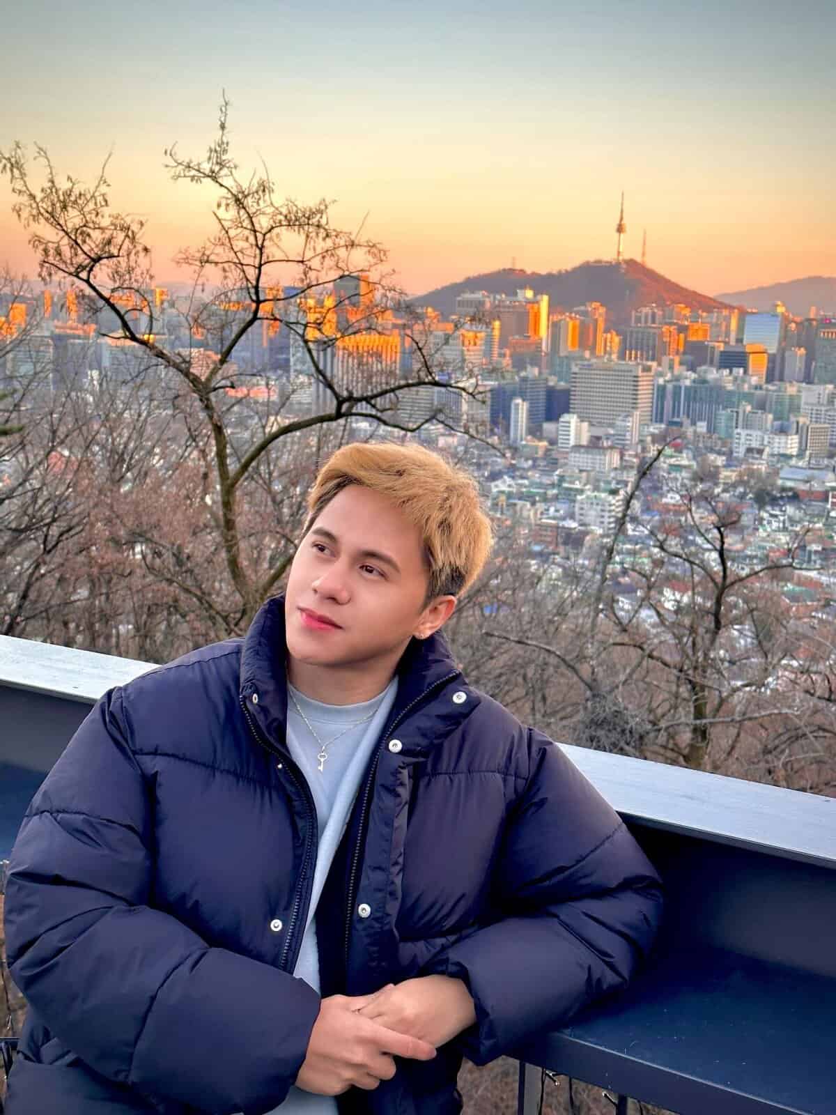 Pinoy scientist Jayson Pasaol is in a dark puffer jacket and leaning on a railing, looking thoughtfully to the side, with a cityscape and a mountain topped with a tower in the background at sunset.