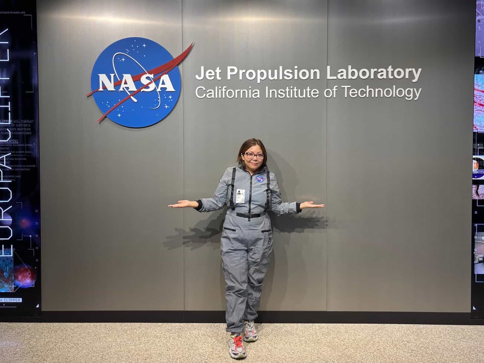 Pinoy scientist Carmela Gonzales in gray suit stands in front of NASA JPL sign, arms out to the sides.