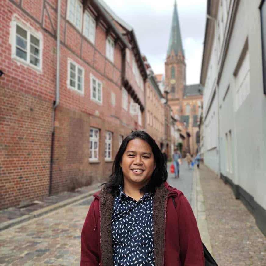 Pinoy scientist Erwin Don Racasa smiling on a cobblestone street with brick buildings and a church in the background.