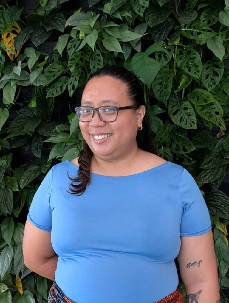 Pinoy scientist Ruby Shaira Panela with glasses and in blue shirt smiling, standing in front of a leafy green wall.