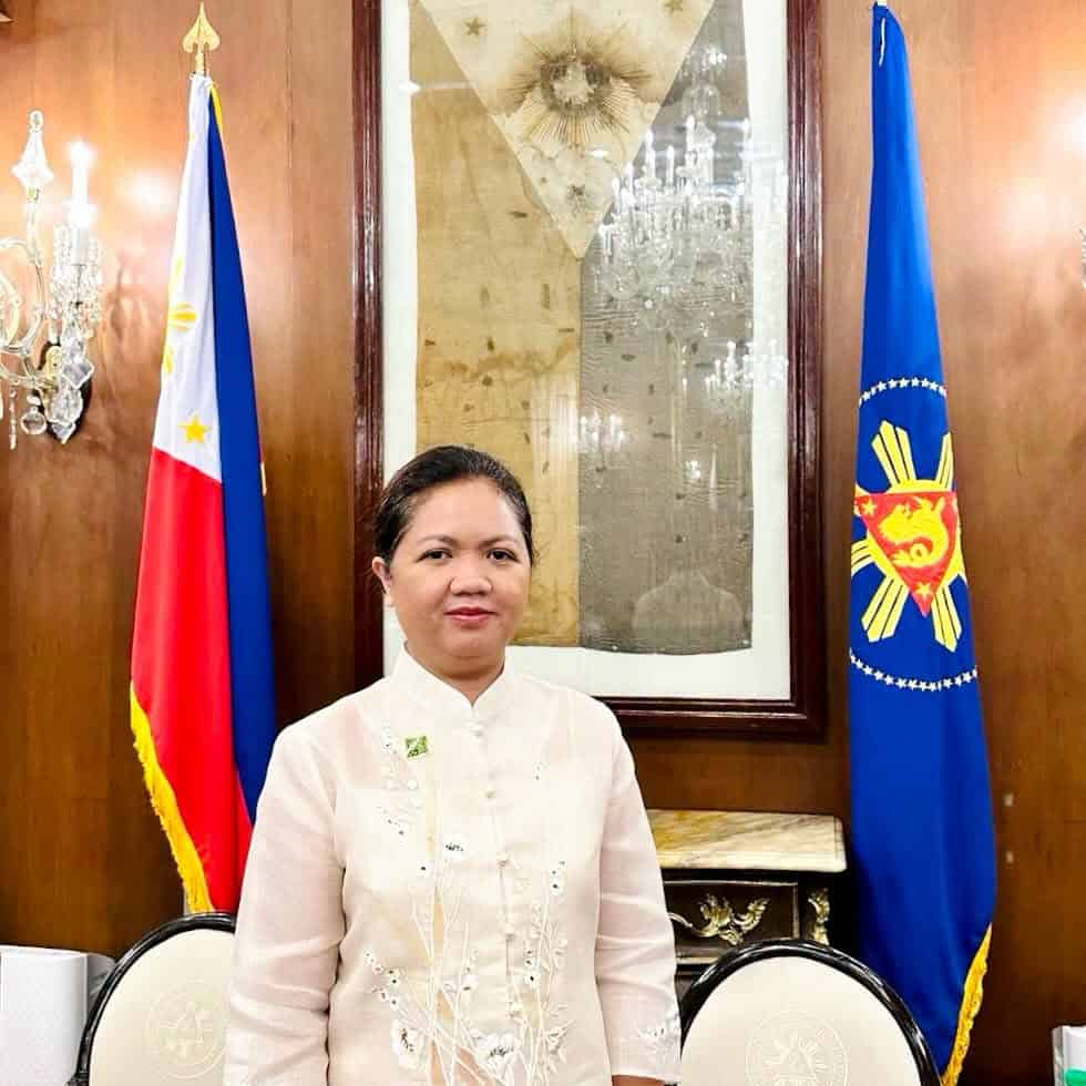 Pinoy scientist Dr. Gay Jane Perez stands between a Philippine flag and a presidential seal in a formal room with wooden walls
