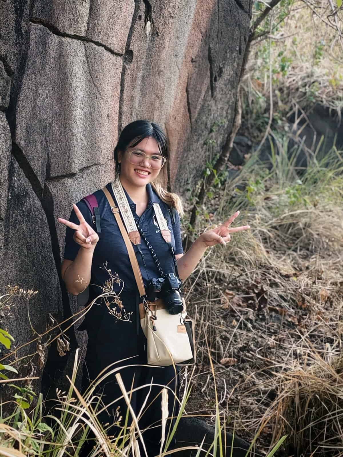 Pinoy scientist Camille Duran with cameras poses, smiling, and making peace signs near a rock wall outdoors.